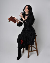 Fototapeta premium Full length portrait of dark haired woman wearing black victorian witch costume. sitting pose on a chair, with gestural hand movements, against studio background.