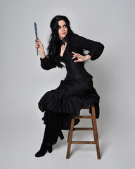 Full length portrait of dark haired woman wearing  black victorian witch costume.  sitting pose on a chair, with gestural hand movements,  against studio background.