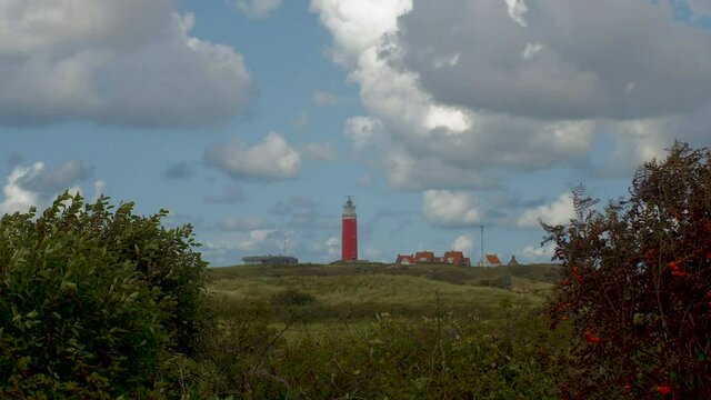 Time lapse through bushes in the foreground. In the distance we see a lighthouse in the dunes together with some builings. The sky is blue with beautiful clouds passing by.