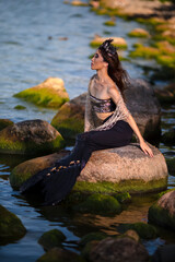 Dreaming Asian Mermaid With Net At Sea Coast on Rocks While Wearing Seashell Decorated Crown and Black Shiny Tail On Sexy Body Covered With Strasses