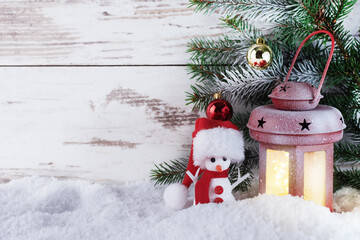 Christmas lantern on snow with fir branch and snowman in a Santa hat over wooden background. Christmas greeting card.