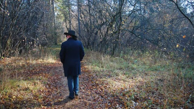 A man in a black hat and a dark coat is walking along a forest path. He is resting outside the city on a day off. The view of a person from the back. Bushes and trees grow along the edge of the path.