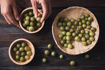 Wild Indian gooseberry or amla in a bamboo basket and bowl with hand on wooden background, Fruit tree in Asia use in various cuisine, herbal medicine and rich vitamin C
