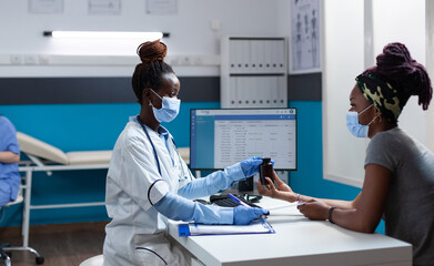 Obraz premium African american doctor with protective face mask against covid19 explaining medication treatment to sick patient holding pills bottle during clinical examination. Physician working in hospital office