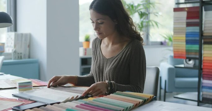 Professional decorator sitting at desk in her studio and choosing fabric swatches, she is touching the samples and checking the material