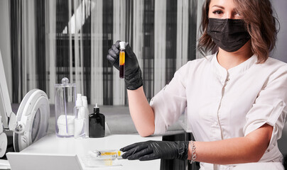 Cosmetologist wearing black gloves, face mask and white uniform, sitting at the table holding blood...