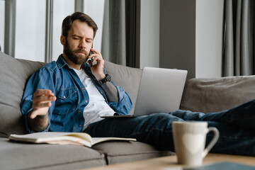 Bearded man talking on cellphone and using laptop while working at home