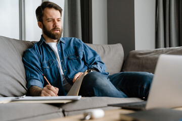 Bearded european man writing down notes while sitting on sofa