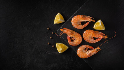 boiled shrimps lie on a stone board on a black concrete surface with lemon wedges and peas. top view. dark photo with copy space