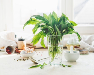 Wild garlic bunch in glass at kitchen table with mortar and pestle, bowls, spices and spoon at...