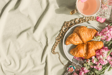 Croissant breakfast background on pale tablecloth with pink tea in tea cup, gold chain and pink flowers. Romantic french sweet breakfast at home. Top view with copy space.