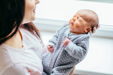 Mother holding cute newborn baby boy in a tender embrace of her arms. Sleepless baby with open eyes funny face expression looking at everything around.