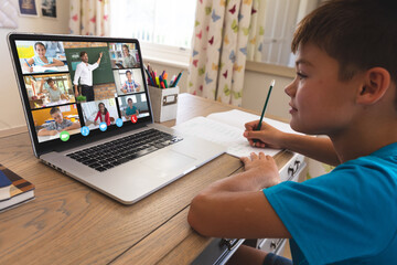 Caucasian boy using laptop for video call, with smiling diverse elementary school pupils on screen