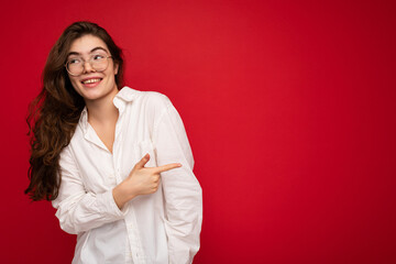 Beautiful happy smiling brunette woman wearing white shirt and optical glasses. Sexy carefree female person posing isolated near red wall in studio with free space. Positive model with natural makeup