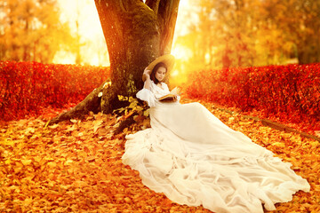 Autumn Landscape. Romantic Victorian Style Woman reading Book in Park with Orange Maple Leaves....