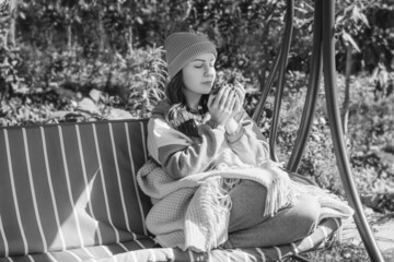 Girl on a swing bench hold autumn drink cup. Autumn day female sit on a swing bench in garden. Gray...