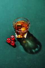 Hard strong alcoholic drinks in glass and casino dice: cognac, tequila, scotch, brandy or whiskey on a green background with hard lights and shadows, top view.