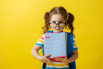 portrait of a cute little girl with glasses in a striped T-shirt with notebooks and textbooks in her hands and a backpack. concept of education. photo studio, yellow background, space for text. 