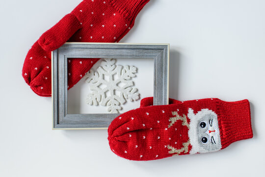 A pair of red woolen knitted soft mittens with cat faces and deer antlers holding a snowflake in a wooden frame on a white background. Christmas and New Years concept. Flat lay, top view.
