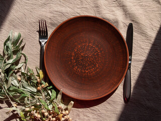 Empty craft handmade clay plate with fork knife linen tablecloth. Table place hand crafted dish. Natural cottagecore styled tableware minimal home interior decor. Restaurant food countryside aesthetic