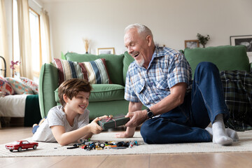 Loving elderly grandfather giving present to smiling grandson. Senior man making surprise to kid boy holding gift box sitting on floor. Excited child receiving congratulations from caring grandpa - obrazy, fototapety, plakaty