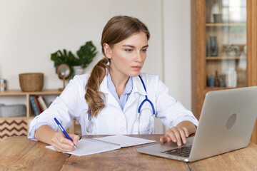 Young caucasian female doctor in white medical uniform working on laptop taking notes at workplace. Woman medic therapist GP fill patient document, writing in journal medical history or anamnesis - obrazy, fototapety, plakaty