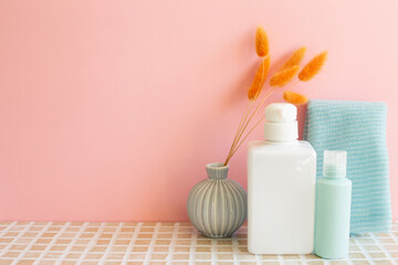 Fototapeta na wymiar Bathroom bottles, shower towel, vase of plant on mosaic tile table. pink wall background. Skin care and spa concept. Home interior