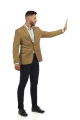 Elegant man is standing with hand raised and showing stop gesture. Side view. Full length isolated.