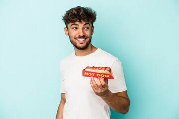 Young arab man holding a hotdog isolated on blue background looks aside smiling, cheerful and...
