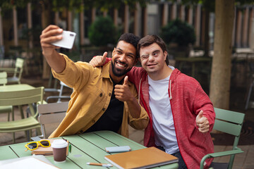 Young man with Down syndrome and his mentoring friend sitting and taking selfie outdoors in cafe