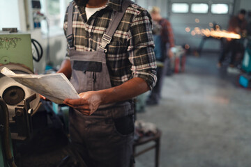 Close up of young industrial man holding blueprints indoors in metal workshop.