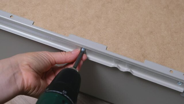 Caucasian woman fastens bolt or lag screw of cabinet drawer slide with electric powered drill. Assembling furniture at home with screw gun