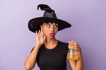 Young mixed race woman disguised as a witch holding potion isolated on purple background  trying to listening a gossip.