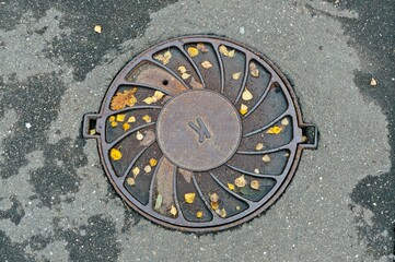 The sewer manhole on the asphalt is covered with autumn leaves
