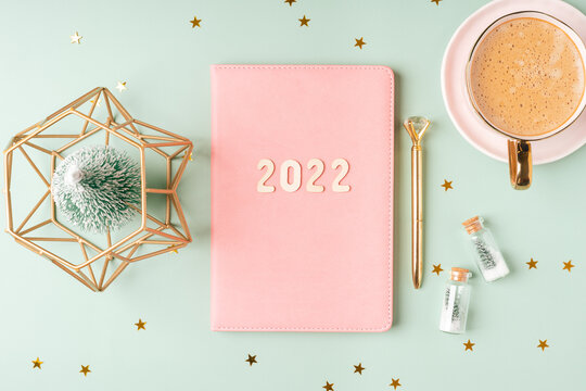 Top view Desktop Christmas pink notepad with 2022 letters text. Flat lay on green mint table background with planner, cup of coffee, candle, Christmas decoration, notebook and stationery. - Powered by Adobe