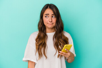 Young caucasian woman holding a mobile phone isolated on blue background confused, feels doubtful...