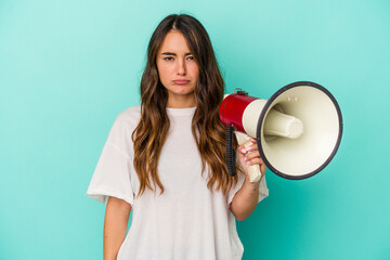 Young caucasian woman holding a megaphone isolated on blue background shrugs shoulders and open eyes confused.