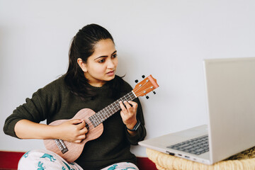 Young woman intently listening to an online class of learning to play Ukulele at home. Laptop...