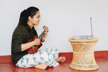 Attractive young woman playing ukulele in front of a laptop. Online learning, teaching, performing,...