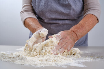 Female hands of an elderly woman knead the dough on a gray table.  Senior woman in a gray apron preparation of dough for a pizza, bread, pasta, festive cake. Shallow depth of field