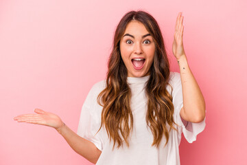 Young caucasian woman isolated on pink background holds copy space on a palm, keep hand over cheek. Amazed and delighted.