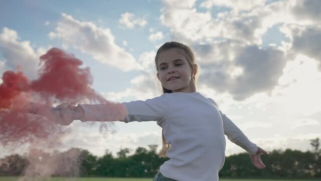 Happy girl dancing outdoors with colorful smoke grenade at sunset. Girl is having fun, waving red smoke bomb in a countryside in slow motion