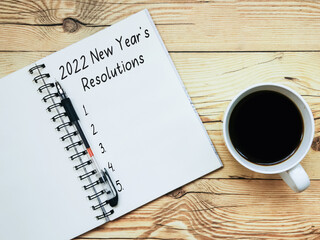 Open notebook with text “2022 New Year's Resolutions” and a cup  of coffee on wooden background.