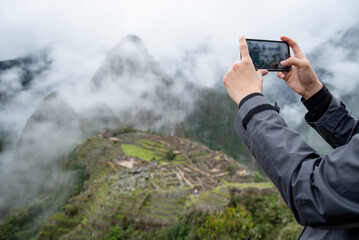 Male tourist taking photo of Machu Picchu on smartphone, one of seven wonders and famous tourist attraction in Cusco Region of Peru. This majestic place has known as 'Lost City of the Incas'