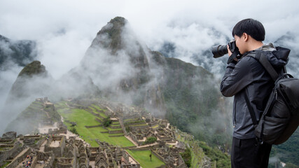 Asian man traveler and photographer taking photo at Machu Picchu, one of seven wonders and famous tourist attraction in Cusco Region of Peru. This majestic place has known as 'Lost City of the Incas'.