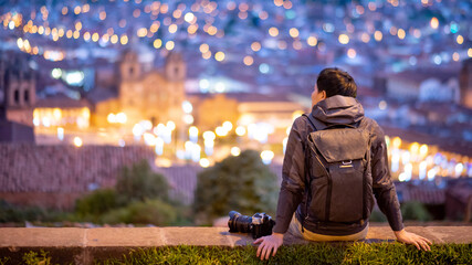 Asian man traveler and photograpaher sitting on viewpoint looking at illuminated cusco city at night. Cusco (Cuzco) is a city in southeastern Peru, near the Urubamba Valley of the Andes mountain range