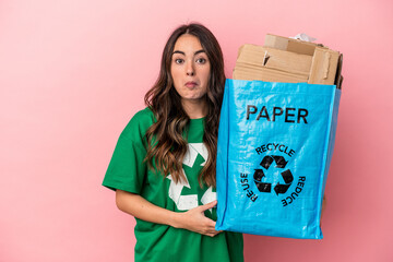 Young caucasian woman recycled paper isolated on pink background shrugs shoulders and open eyes...