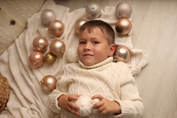 Cute blond boy in a winter sweater is lying on the floor around Christmas balls, the boy is smiling