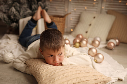 Cute blond boy in a winter sweater is lying on the floor on a pillow around Christmas balls, the boy is sad