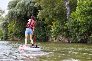 Caucasian woman rowing on stand up paddle board, SUP alone in river near green trees. Back view